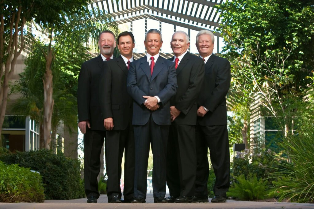 Five men in suits posed outdoors among palm trees
